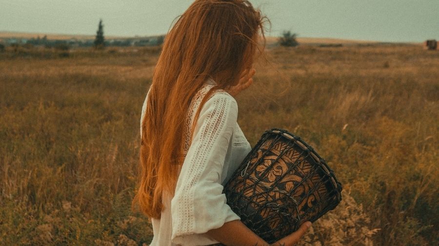 Woman in a white dress walking across a grassy field, carrying a hand drum.