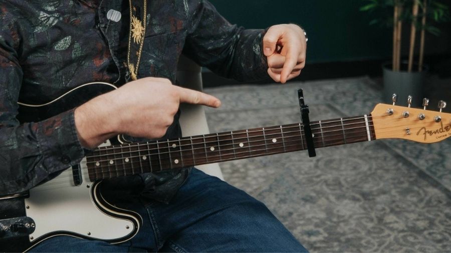 “Man with a guitar on his lap pointing at a guitar accessory.