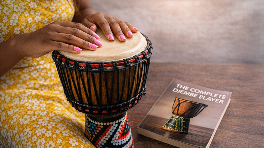 Hands playing a djembe drum beside The Complete Djembe Player book, showing proper hand technique on a traditional African drum in a clean studio setting