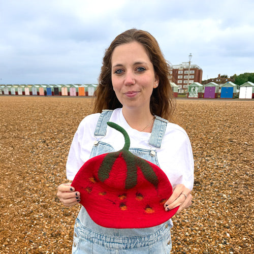 Woman holding a red felt strawberry on a beach with colorful buildings in the background