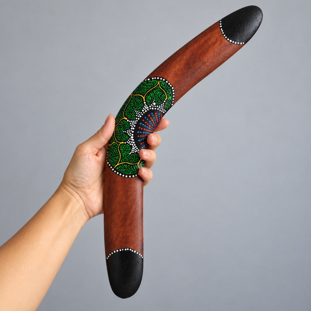 A hand holds a curved wooden boomerang with black-tipped ends and a green, blue, and yellow beaded dot design on the front, set against a plain gray background.
