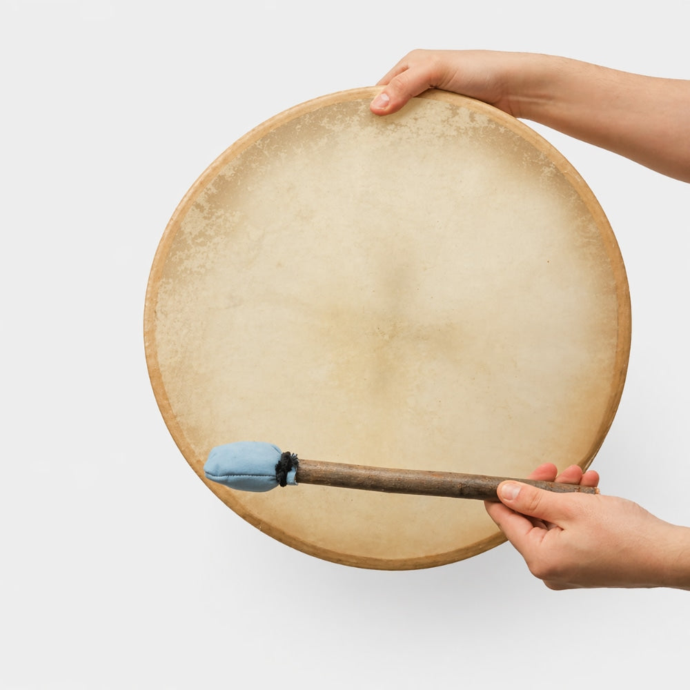 Hand holding large 16" khenji cow hide shamanic drum with a blue tipped beater on a light gray studio background