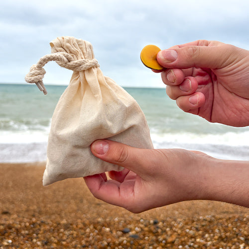 Person holding a small yellow tagua seed above a beige drawstring bag on a beach.