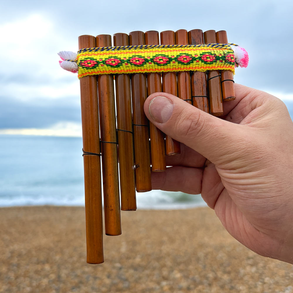 Hand holding a bamboo pan flute with a colorful strap against a beach background