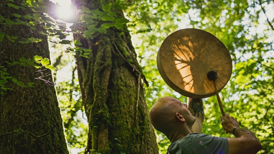 An adult man playing a shamanic drum in a focused, rhythmic manner.