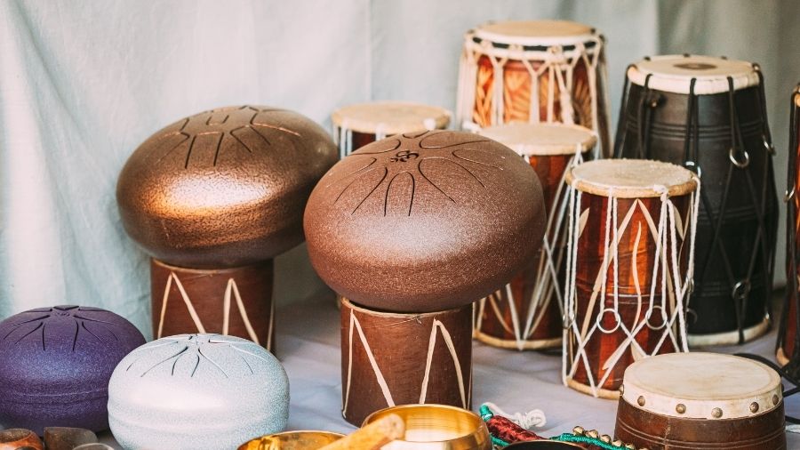 Handmade percussion instruments from Carved Culture including a tongue drum, Indian dhol drum, and Indian bells displayed together.