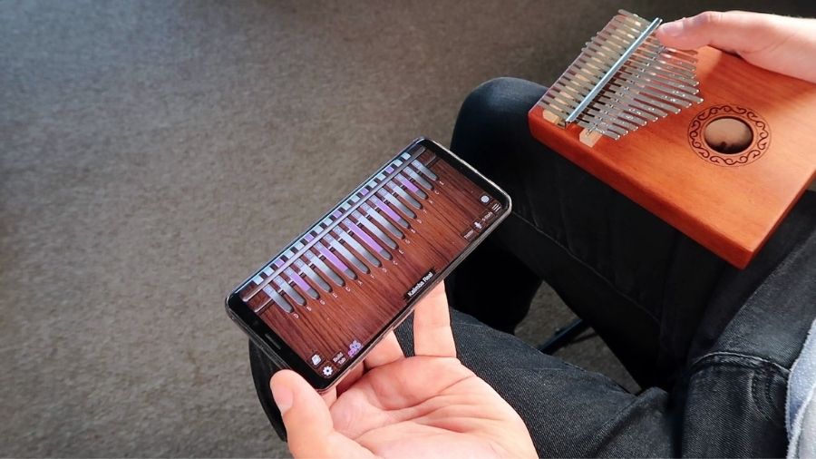 A man holding a kalimba in one hand and a phone showing a kalimba app in the other, comparing them.