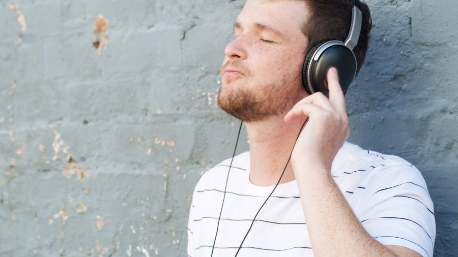 A calm man wearing headphones outdoors, leaning against a gray wall, enjoying music, streaming a zen playlist, or listening to binaural beats for relaxation.