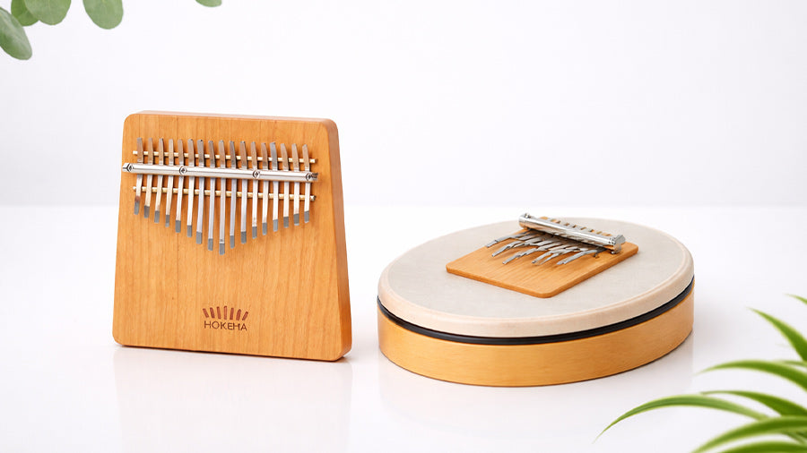 Two Hokema instruments displayed side by side on a white reflective background, showing a wooden kalimba and a Sansula kalimba with natural eucalyptus and spider plant leaves in a minimalist studio setup.