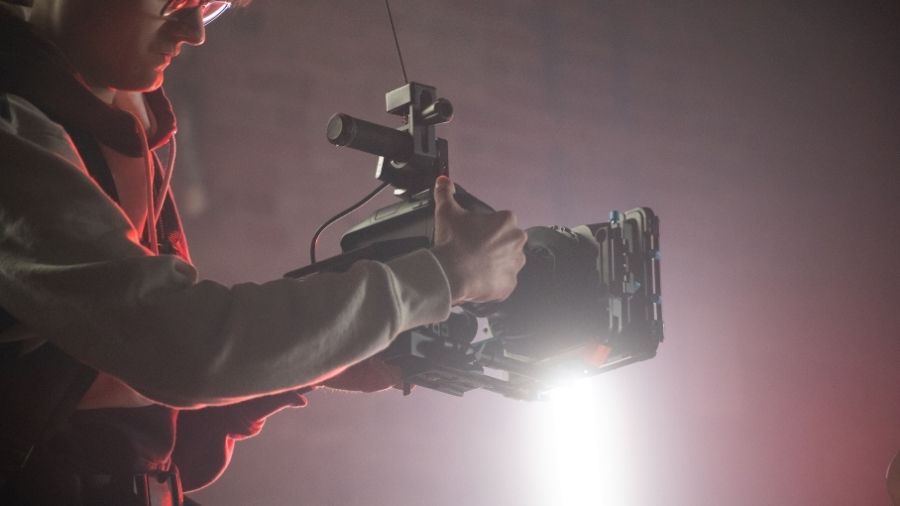 Cinematographer filming a music video with a camera, soft pink backdrop, and stage lighting.
