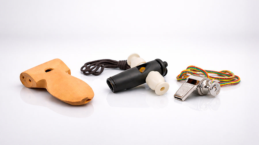 Three samba whistles displayed in a minimalist studio setting, including a wooden apito whistle, a black and white plastic tritone whistle, and a metal samba whistle with a rainbow cord.