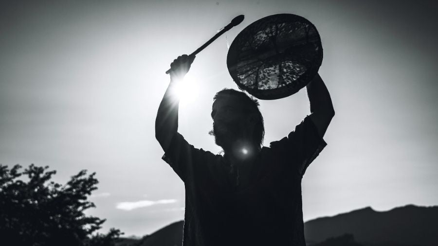 Silhouette of a shaman standing in the mountains, holding a shamanic drum and drum beater.