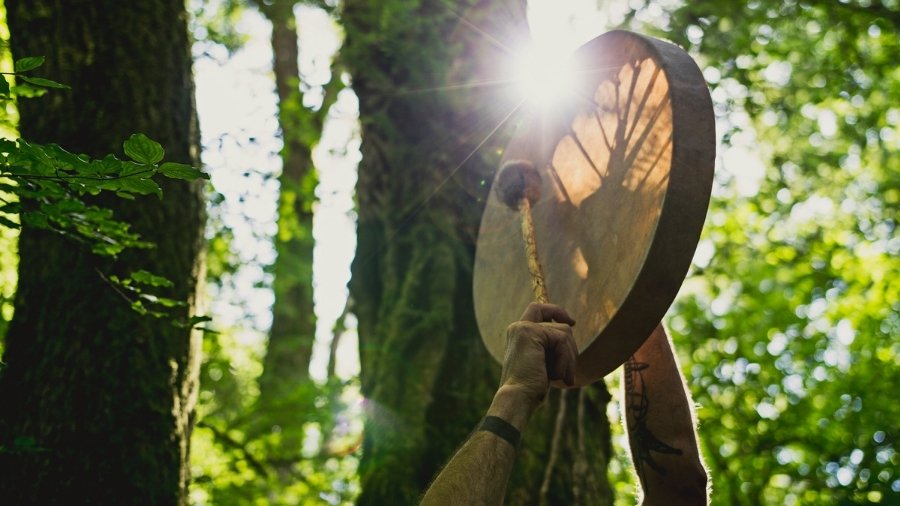 Man holding up a shamanic rattle and drum on a forest background
