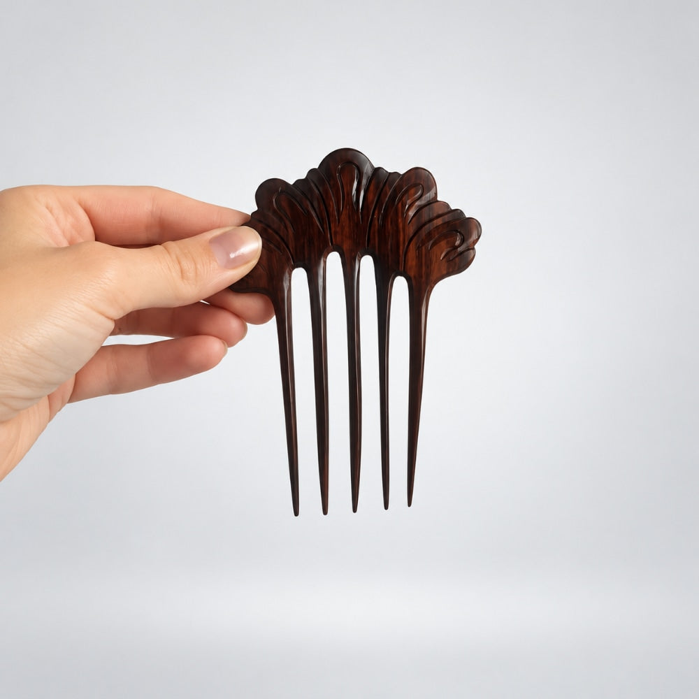 A hand holding a dark brown carved wooden hair comb with five tines against a light gray background.