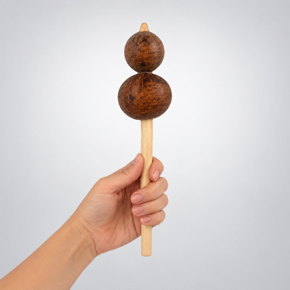 A hand holding a wooden double-gourd rattle against a light gray background.