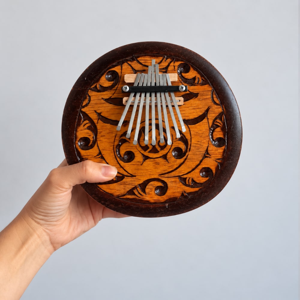 A hand holding a round carved wooden kalimba thumb piano against a plain light gray background.