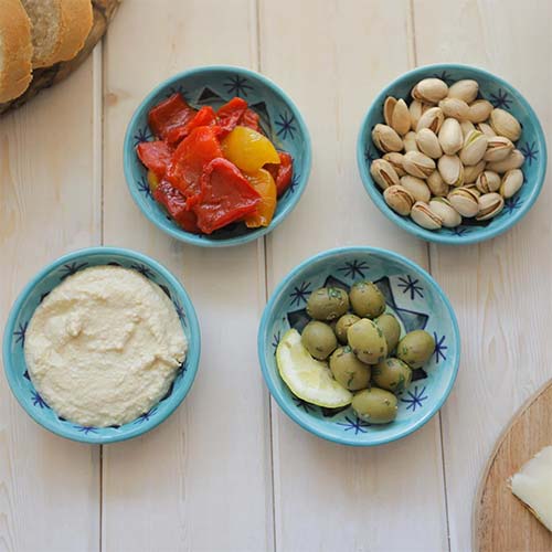 A set of four small tapas Patna bowls with lemon herb olives, hummus, roasted red peppers, pistachios on a rustic white washed wood background 
