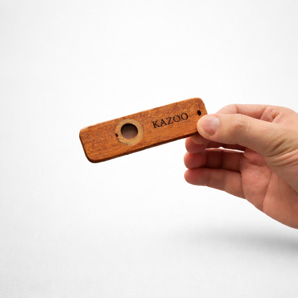 Hand holding a wooden kazoo instrument to show scale with 'KAZOO' branding on a white background.